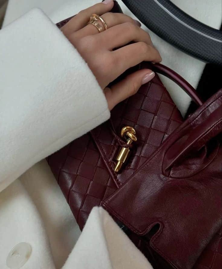Burgundy Winter Details Hand with gold rings resting on a stylish maroon woven leather bag, draped over a chic white coat. | Sky Rye Design Hand with gold rings resting on a stylish maroon woven leather bag, draped over a chic white coat.
