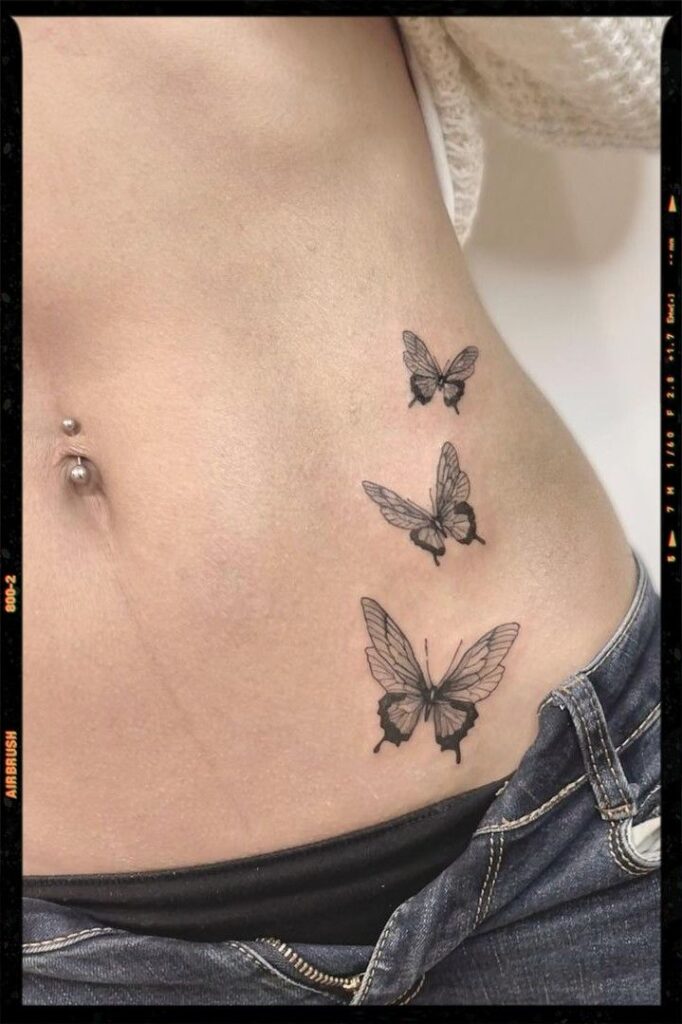 Butterfly Tattoos Butterfly tattoos on a person's side with a belly button piercing, showcasing delicate artistry in black ink above denim jeans. | Sky Rye Design Butterfly tattoos on a person's side with a belly button piercing, showcasing delicate artistry in black ink above denim jeans.