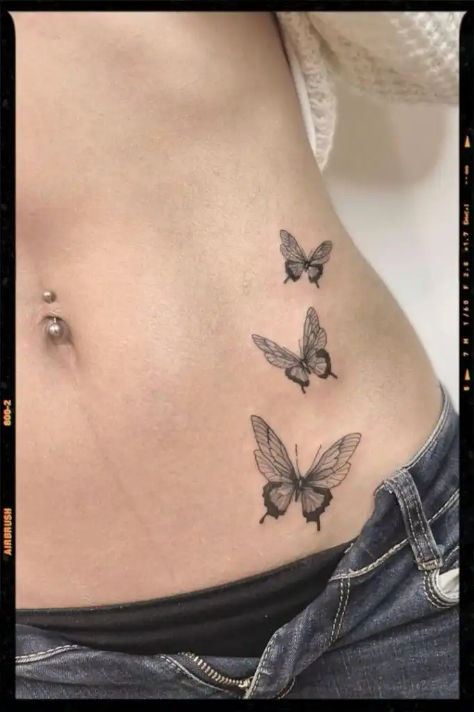 Butterfly Tattoos Butterfly tattoos on a person's side with a belly button piercing, showcasing delicate artistry in black ink above denim jeans. | Sky Rye Design Butterfly tattoos on a person's side with a belly button piercing, showcasing delicate artistry in black ink above denim jeans.