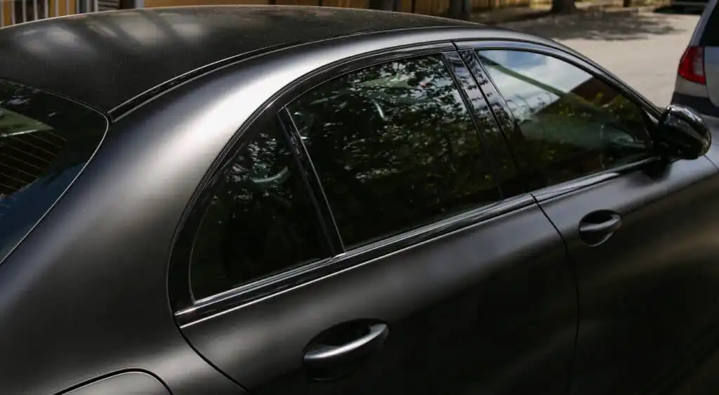 Sleek black sedan in sunlight, showing smooth curves and tinted windows, parked on a quiet street with reflections of nearby trees.