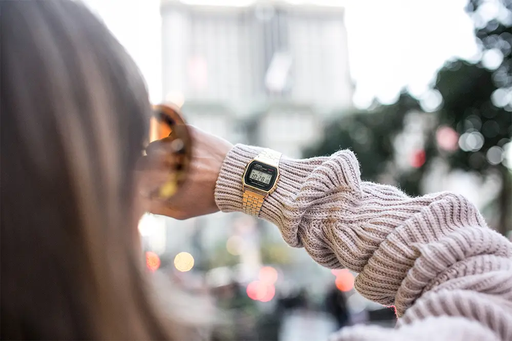 Woman wearing vintage digital gold watch, cityscape blurred in background, focusing on timepiece for stylish urban fashion statement.