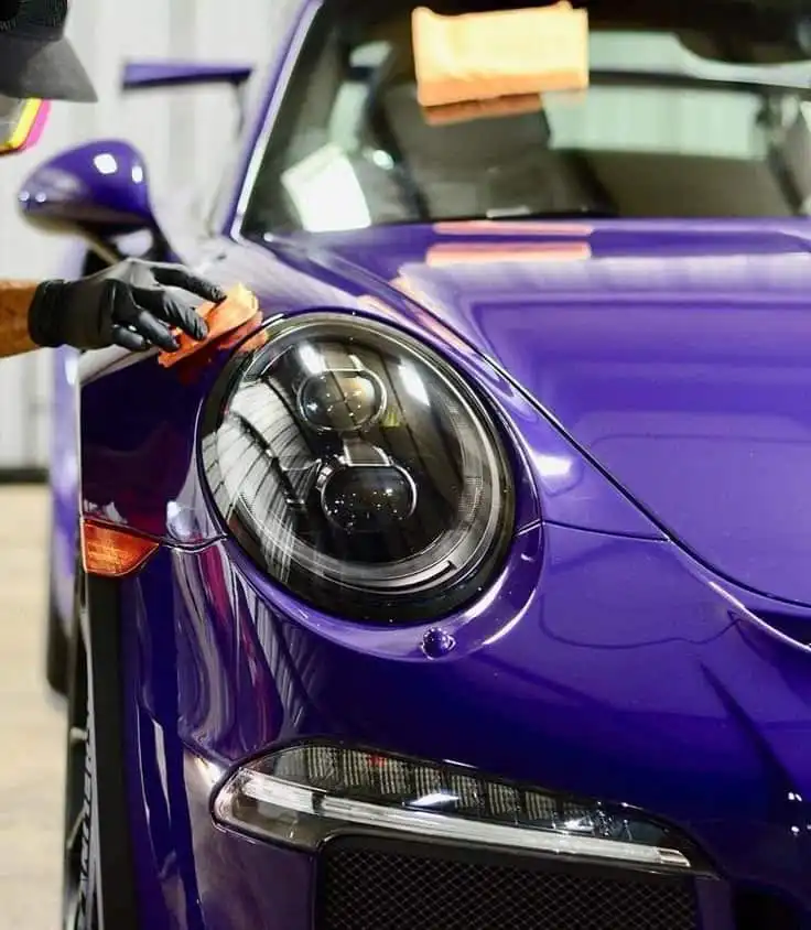 Close-up of a person polishing the headlight of a shiny purple sports car, highlighting sleek design and car detailing.