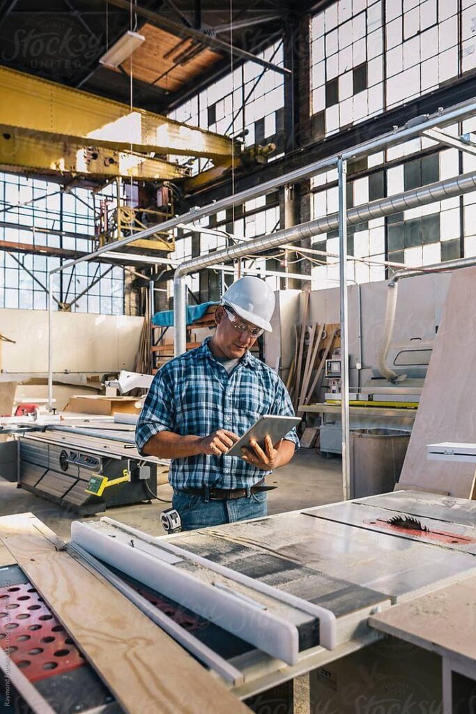 _Engineer Using Laptop Computer_ by Stocksy Contributor _Raymond Forbes LLC_ Man in a hard hat using a tablet in a woodworking shop, surrounded by tools and equipment, under natural light from large windows. | Sky Rye Design Man in a hard hat using a tablet in a woodworking shop, surrounded by tools and equipment, under natural light from large windows.