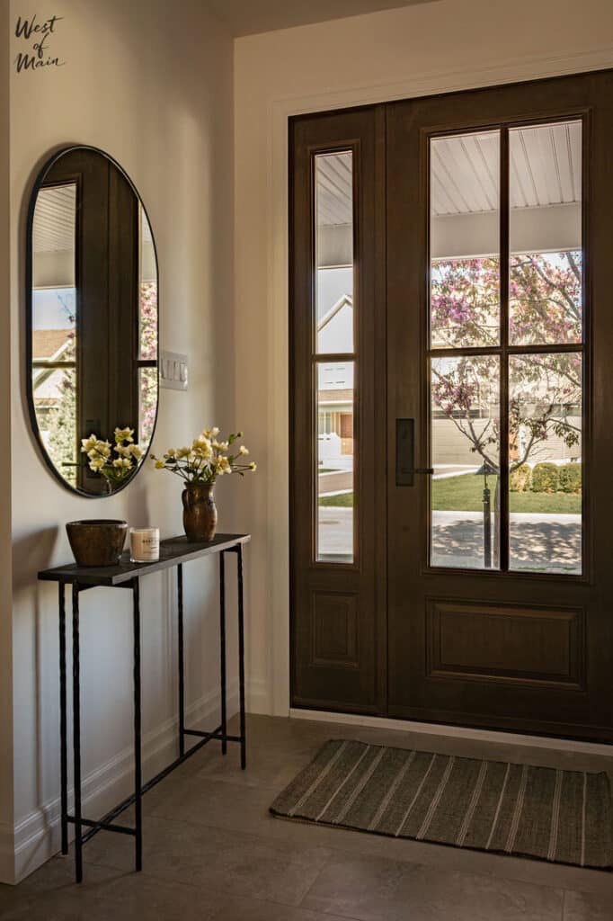 Entryway Round Up _ West of Main Modern entryway with wooden door, oval mirror, minimalist console table, and fresh flowers. Bright, welcoming interior design. | Sky Rye Design Modern entryway with wooden door, oval mirror, minimalist console table, and fresh flowers. Bright, welcoming interior design.