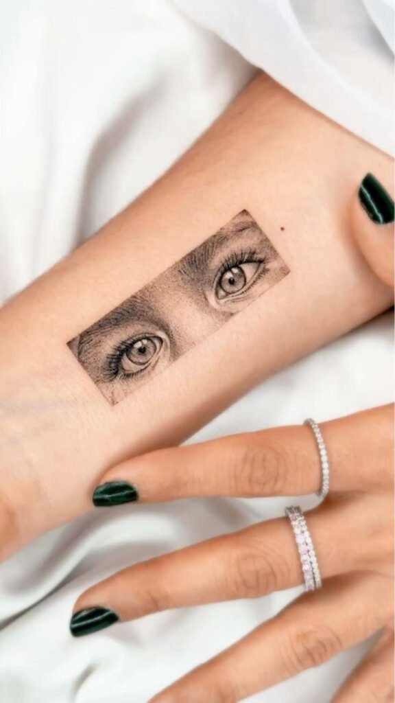Eyes _ Small Tattoo Realistic eye tattoo on forearm with detailed shading, surrounded by elegant hands wearing rings, featuring dark green nail polish. | Sky Rye Design Realistic eye tattoo on forearm with detailed shading, surrounded by elegant hands wearing rings, featuring dark green nail polish.