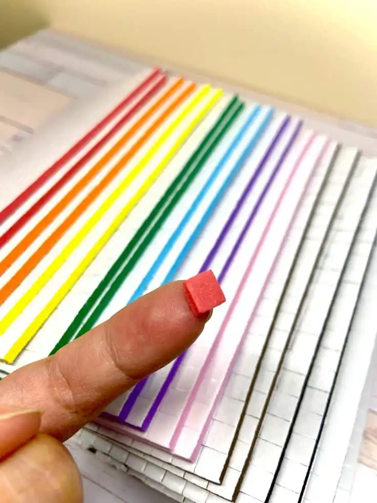 Tiny red cube on fingertip, with colorful, aligned paper strips creating a rainbow effect in the background.
