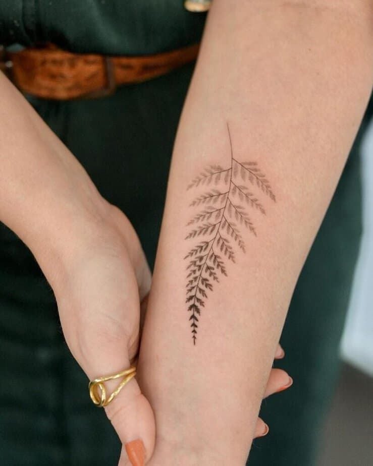 Delicate fern tattoo on forearm with gold ring, showcasing a minimalist botanical design for nature-inspired body art enthusiasts.