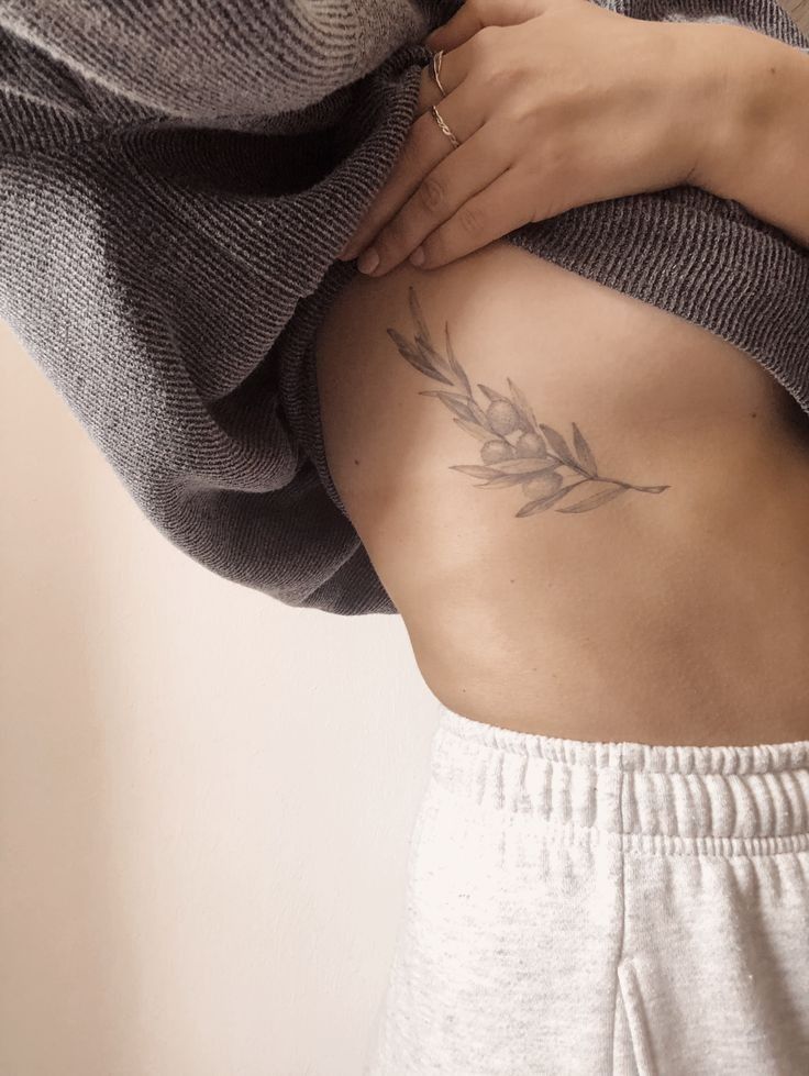 Woman showcasing a delicate olive branch tattoo on her side, wearing a cozy gray sweater and light sweatpants for a casual look.