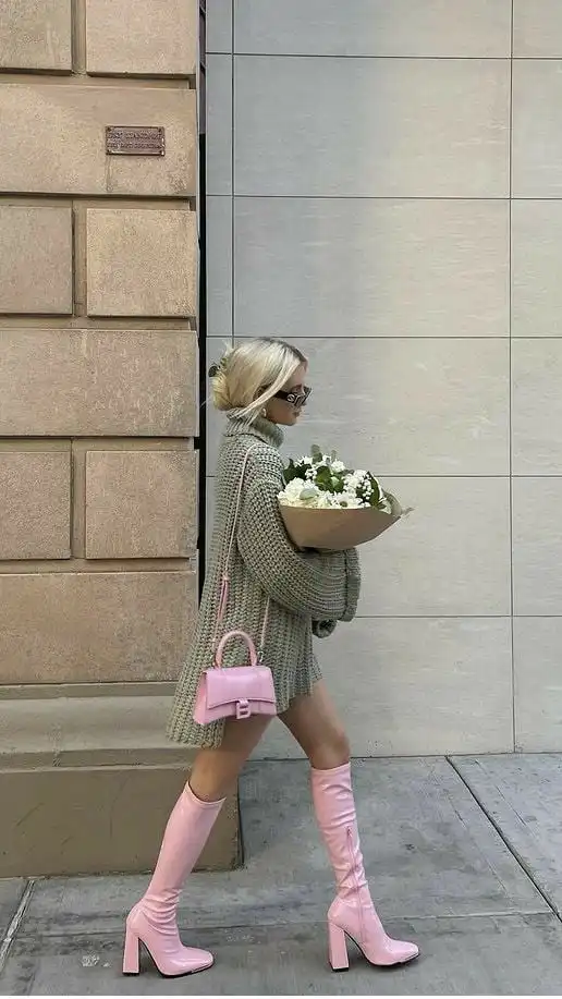 Woman in a gray sweater, pink boots, and sunglasses walking with a pink bag and bouquet on a city sidewalk. Fashion street style.