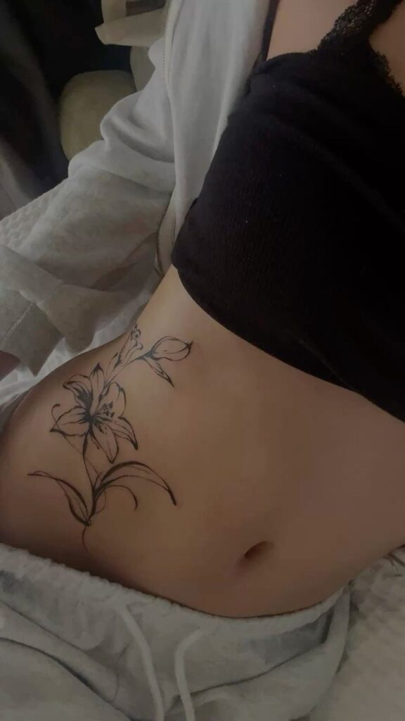 Floral waist_stomach tattoo Floral side tattoo on a woman's stomach, featuring a delicate lily, styled with a casual black crop top and white robe. | Sky Rye Design Floral side tattoo on a woman's stomach, featuring a delicate lily, styled with a casual black crop top and white robe.