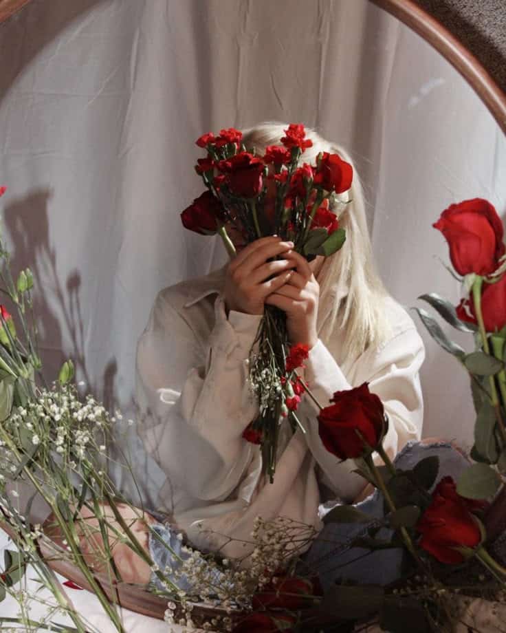 Florals shoot Person holding a bouquet of red roses, seated in front of a mirror, surrounded by flowers, wearing a light-colored shirt. | Sky Rye Design Person holding a bouquet of red roses, seated in front of a mirror, surrounded by flowers, wearing a light-colored shirt.