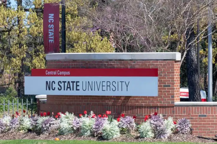 Sign for NC State University Central Campus, surrounded by trees and flowers, under a clear blue sky.