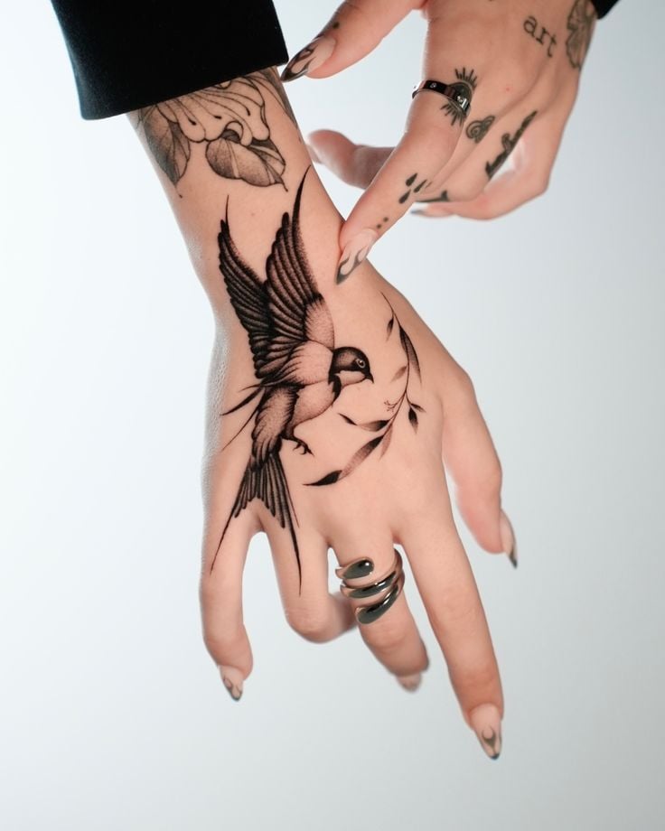 Hand with a detailed bird tattoo and floral design, adorned with rings and nail art on a white background.
