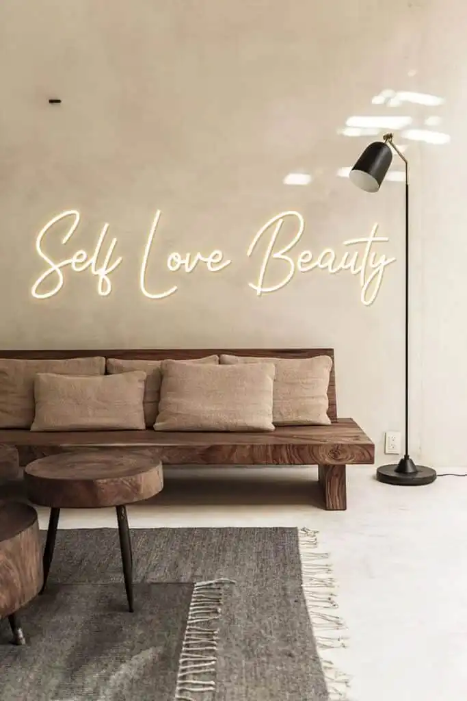 Modern minimalist room with Self Love Beauty neon sign above a cozy wooden sofa, neutral cushions, round tables, and a floor lamp.
