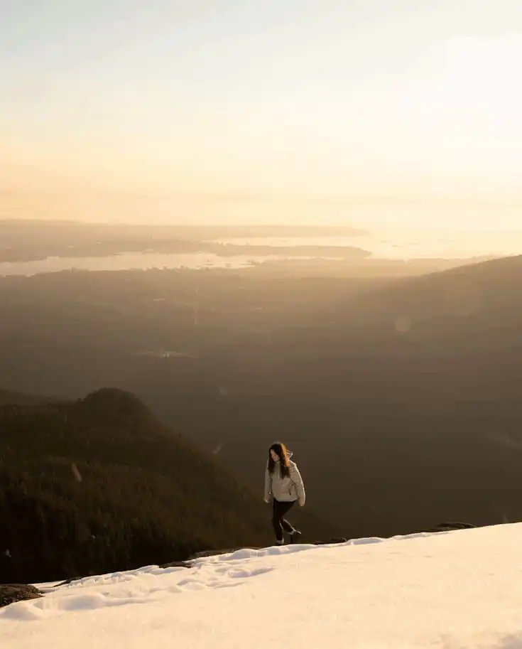 Person hiking in snowy mountains at sunrise, overlooking a vast forest and distant cityscape under a warm golden sky.