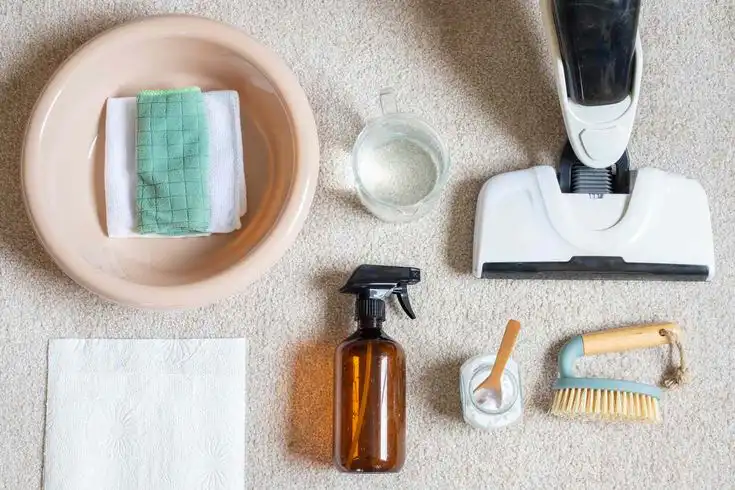 Overhead view of carpet cleaning tools: vacuum, spray bottle, scrub brush, bowl with cloths, and a jar on a beige carpet