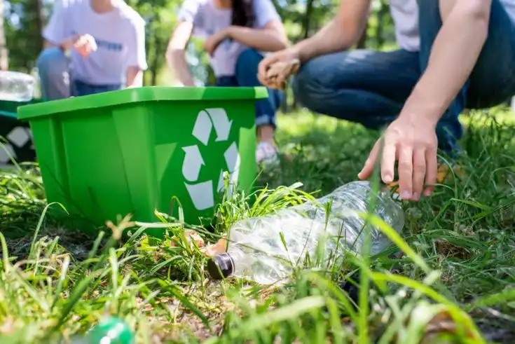 Group of people recycling plastic outdoors, placing bottles in a green bin with a recycling symbol, promoting environmental
