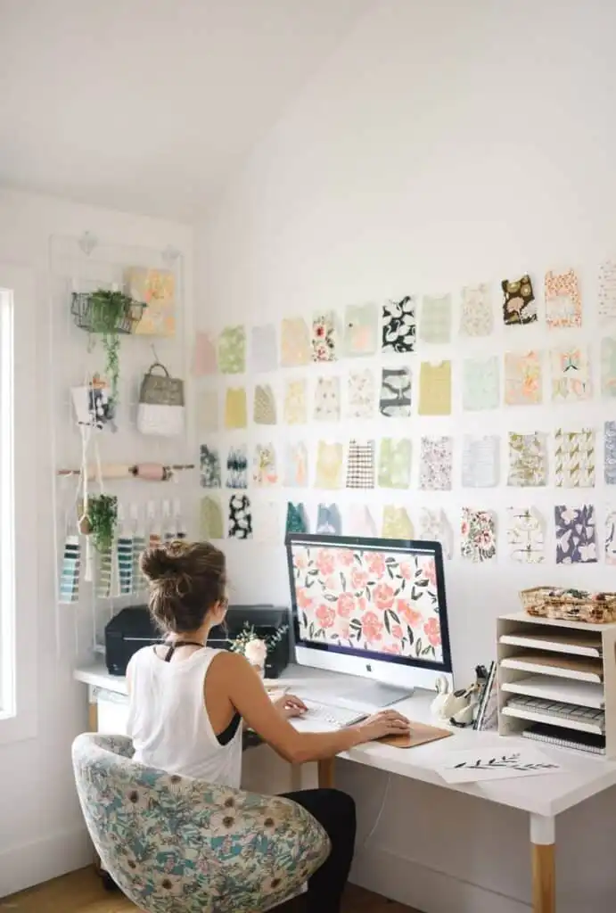 Designer working in a creative studio, surrounded by floral patterns and fabric samples on walls, using a desktop computer.