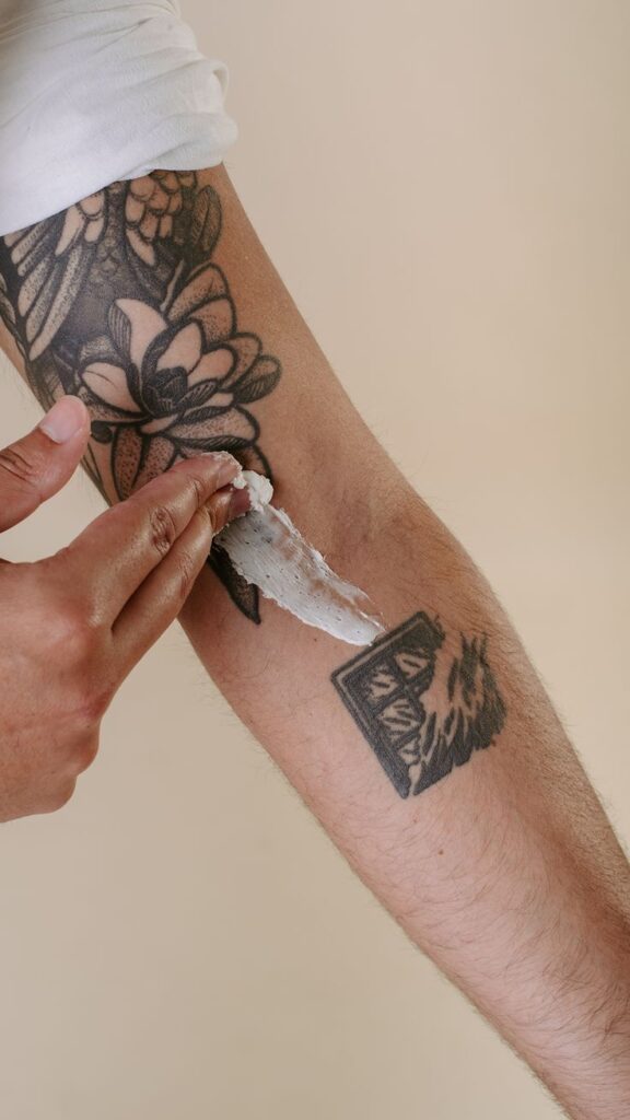 Humecta el Tattoo Person applying cream on tattooed forearm, highlighting proper tattoo care with moisturizing lotion on floral and geometric designs. | Sky Rye Design Person applying cream on tattooed forearm, highlighting proper tattoo care with moisturizing lotion on floral and geometric designs.