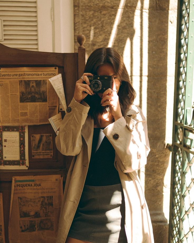 Instagram Woman in a trench coat taking a photo with a vintage camera indoors, surrounded by newspaper clippings and sunlight streaming through. | Sky Rye Design Woman in a trench coat taking a photo with a vintage camera indoors, surrounded by newspaper clippings and sunlight streaming through.