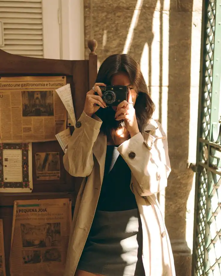 Instagram Woman in a trench coat taking a photo with a vintage camera indoors, surrounded by newspaper clippings and sunlight streaming through. | Sky Rye Design Woman in a trench coat taking a photo with a vintage camera indoors, surrounded by newspaper clippings and sunlight streaming through.