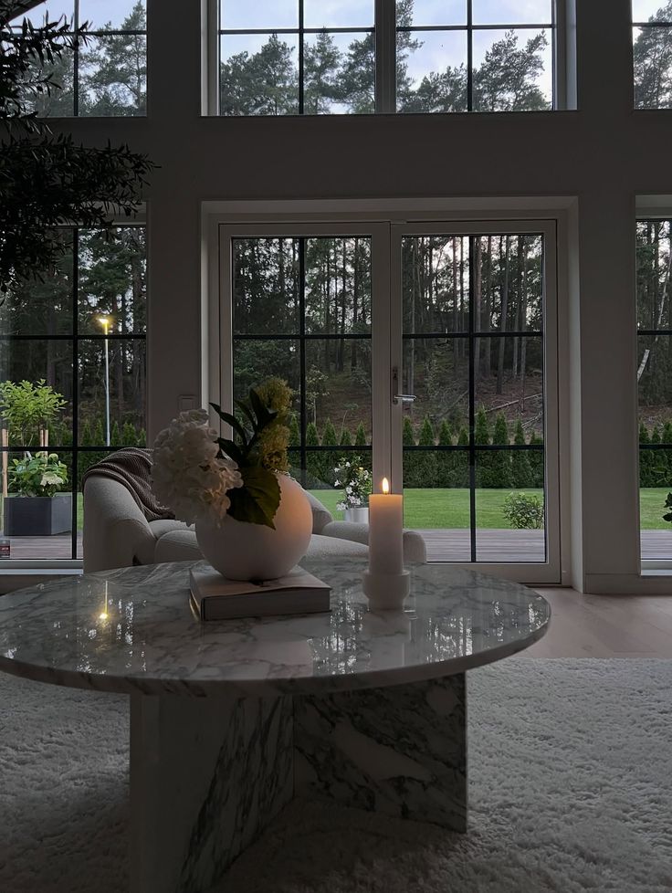 Instagram Sophisticated living room with large windows, marble table, candle, and greenery, offering a serene view of the lush outdoor garden. | Sky Rye Design Sophisticated living room with large windows, marble table, candle, and greenery, offering a serene view of the lush outdoor garden.