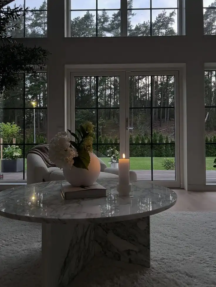 Instagram Sophisticated living room with large windows, marble table, candle, and greenery, offering a serene view of the lush outdoor garden. | Sky Rye Design Sophisticated living room with large windows, marble table, candle, and greenery, offering a serene view of the lush outdoor garden.