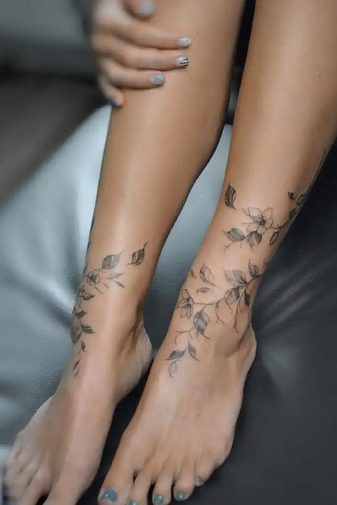Floral ankle tattoos on legs with floral nail art sitting on a leather couch, showcasing delicate and elegant body art.