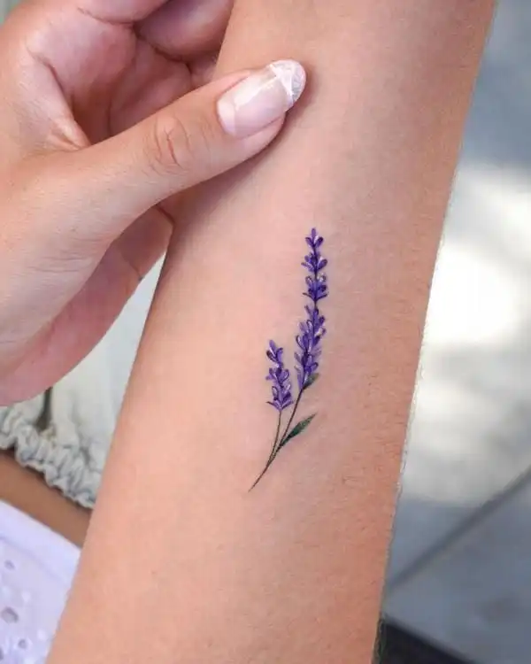Delicate lavender flower tattoo on forearm, held by a hand with manicured nails. Minimalist floral ink design.