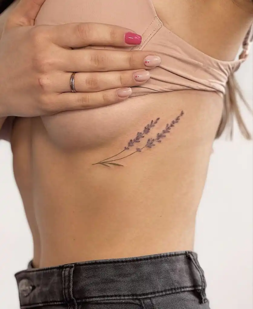 Close-up of a minimalist lavender flower tattoo on a person's side, under a lifted beige top, showcasing pink and nude manicured nails.