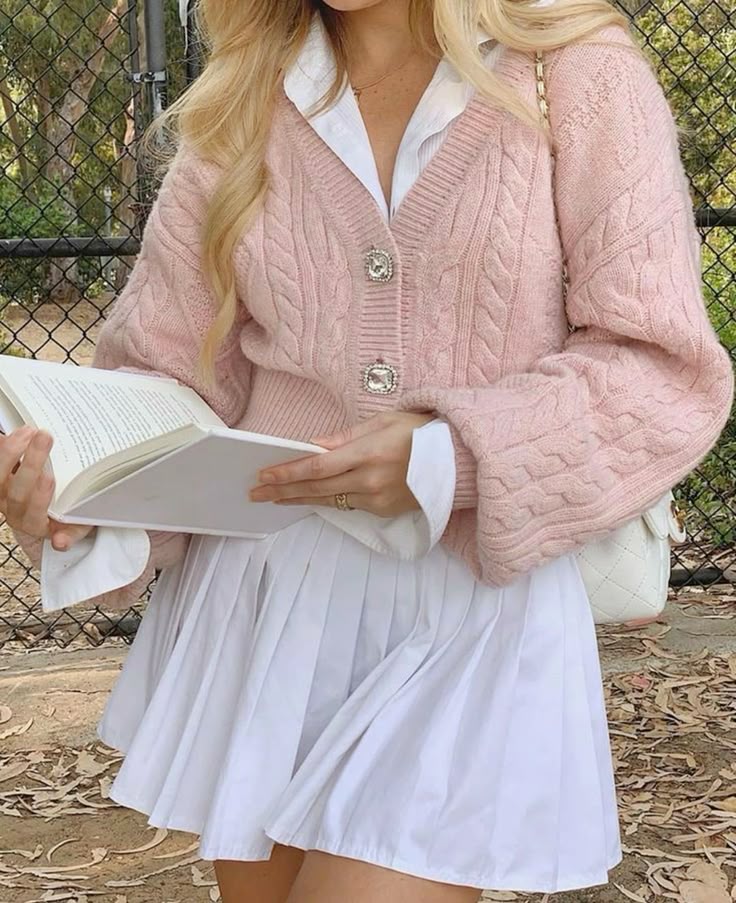 Light Academia Cardigan _ Rusty Pink Woman in pink cable-knit sweater and white dress reading a book outdoors, showcasing casual chic fall fashion style. | Sky Rye Design Woman in pink cable-knit sweater and white dress reading a book outdoors, showcasing casual chic fall fashion style.