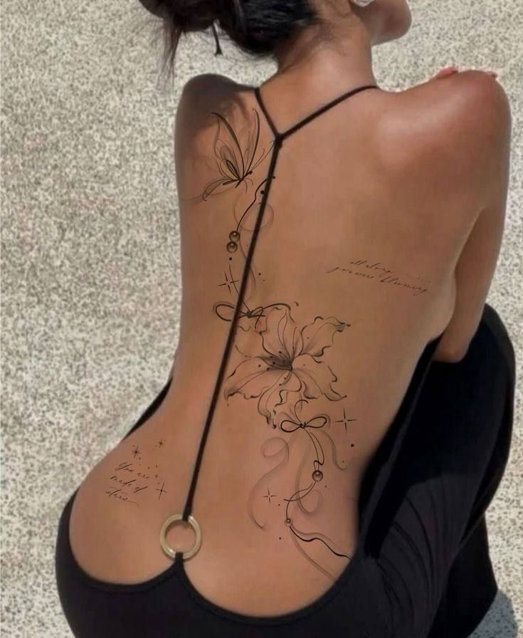 Back tattoo featuring floral and abstract designs on a woman wearing a black strappy dress, sitting outdoors on a textured surface.