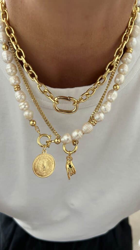 Gold and pearl layered necklaces with charms, including a coin and hand, elegantly styled on a white shirt background.