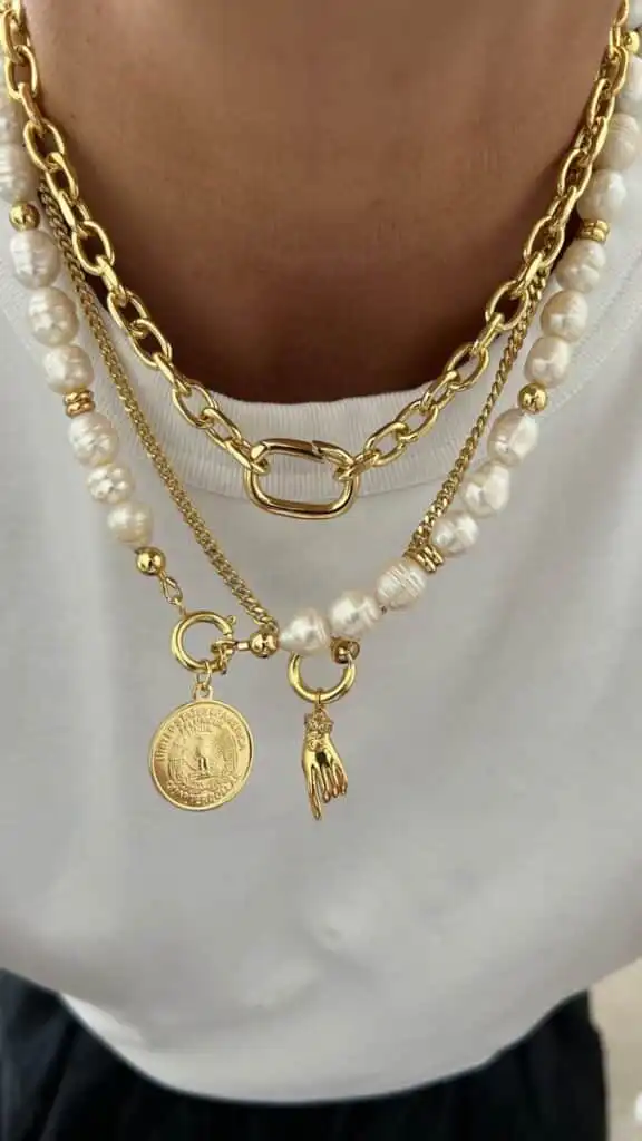 Gold and pearl layered necklaces with charms, including a coin and hand, elegantly styled on a white shirt background.