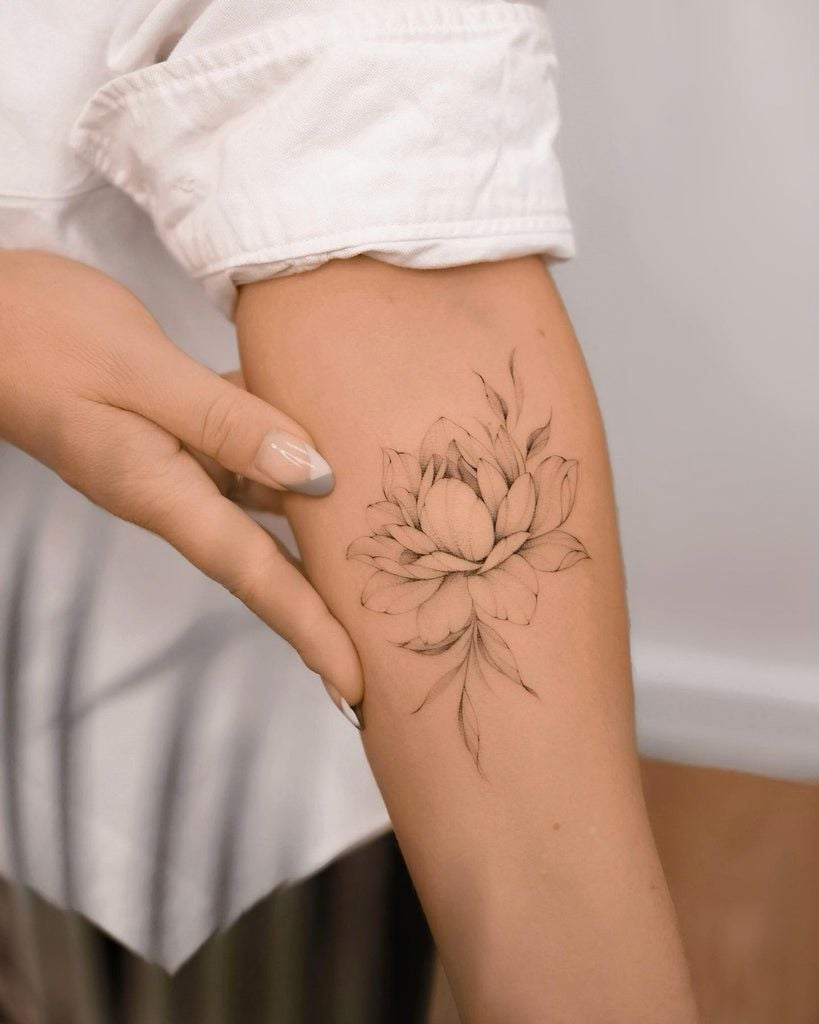 Person showcasing a delicate black floral tattoo on their arm, highlighting intricate design and elegance with a casual white shirt backdrop.