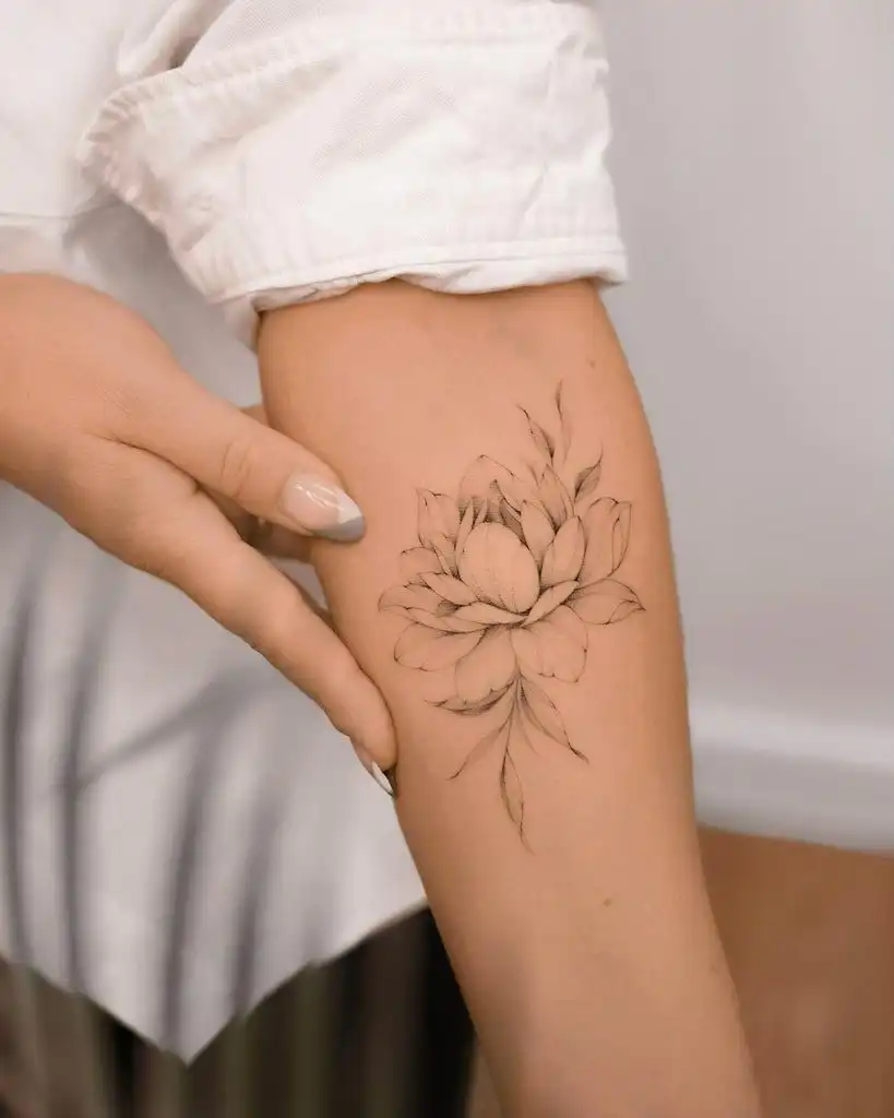 Person showcasing a delicate black floral tattoo on their arm, highlighting intricate design and elegance with a casual white shirt backdrop.