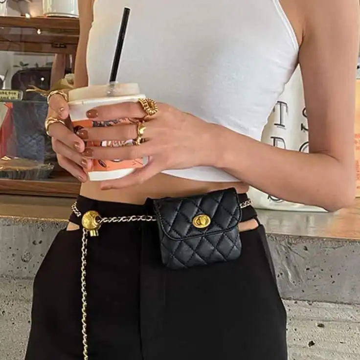 Woman in white crop top holding a drink, wearing gold rings and a quilted black belt bag, showcasing chic fashion accessories.