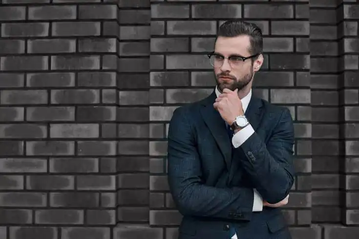 Man in a suit with glasses and a watch, thoughtfully posing against a brick wall, embodying business style and confidence.