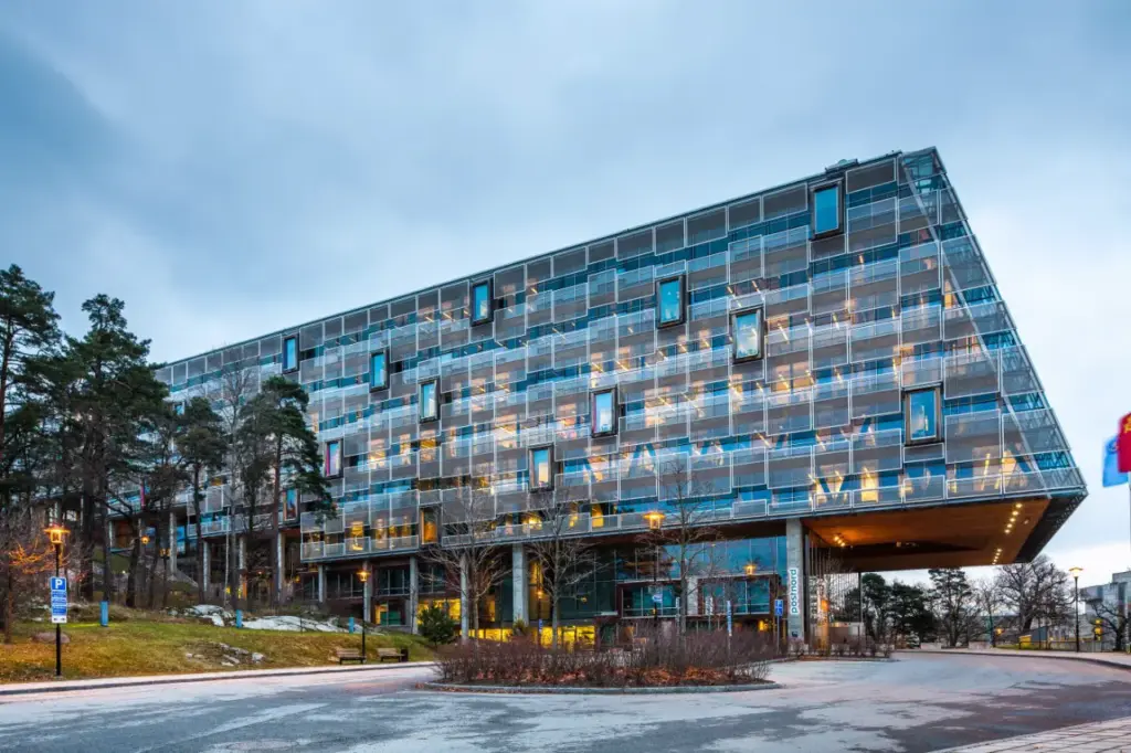 Modern glass office building surrounded by trees and lit by evening lights, showcasing urban architecture against a cloudy sky.