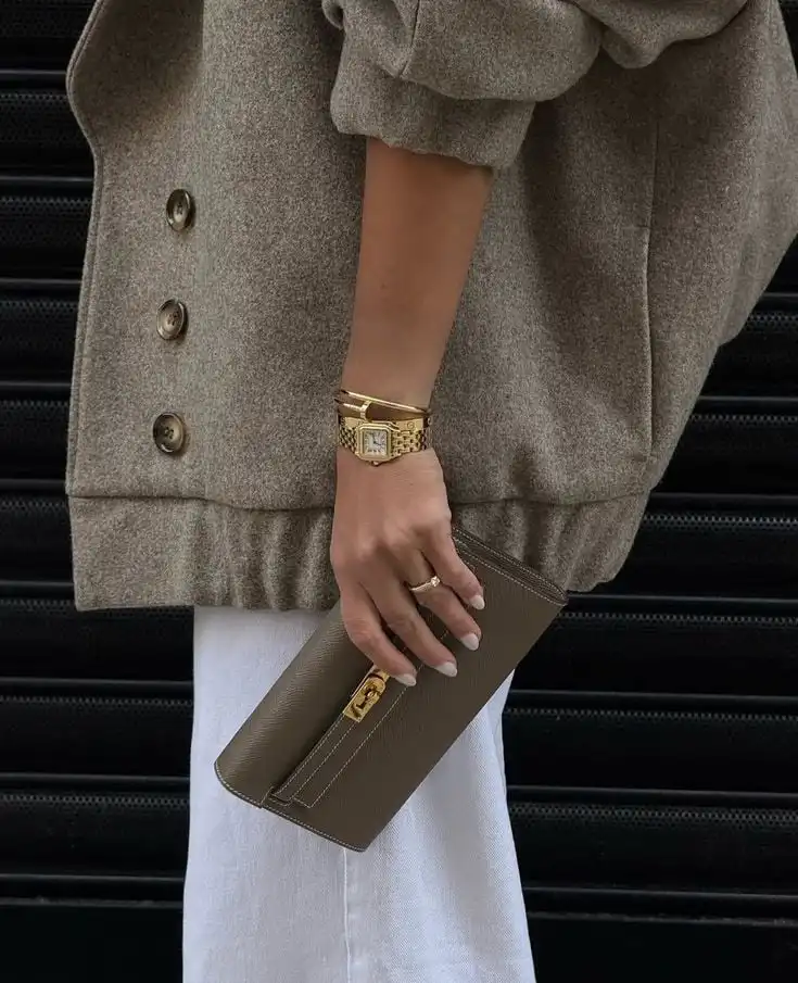 Stylish outfit with beige coat, holding a matching taupe clutch. Gold bracelet watch adds elegance against white pants. Fashion detail shot.