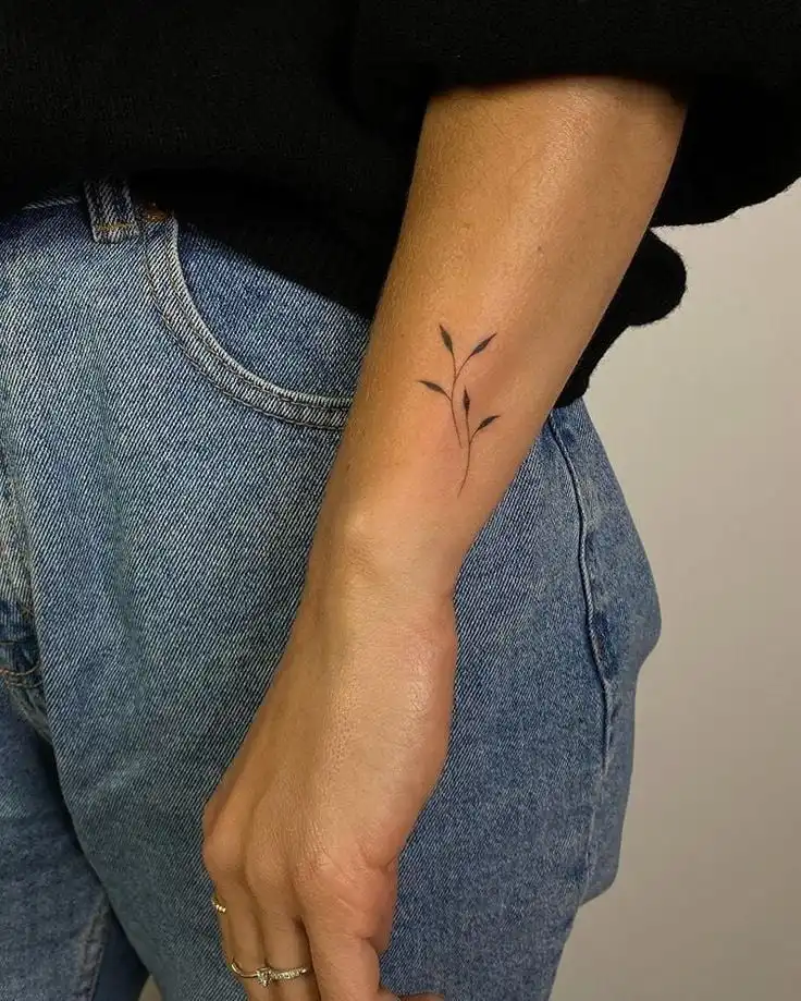 Minimalist leaf tattoo on forearm, wearing black sweater and blue jeans. Simple, elegant design showcases subtle body art style.