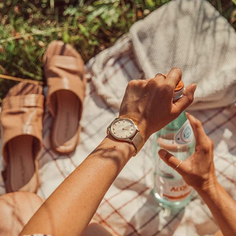 Woman wearing a watch opens a water bottle during a picnic, with sandals and a bag on a plaid blanket, surrounded by grass.