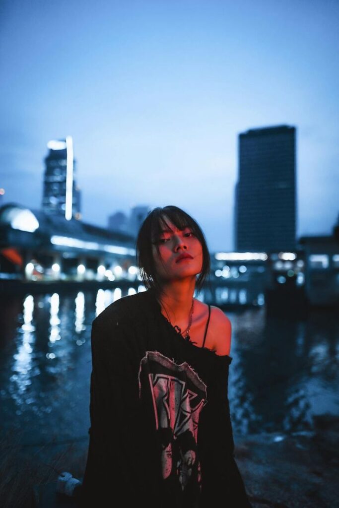 Moody portrait photography Young woman in urban setting at dusk, blue skyline reflected on water. Relaxed, edgy vibe with city lights behind. | Sky Rye Design Young woman in urban setting at dusk, blue skyline reflected on water. Relaxed, edgy vibe with city lights behind.
