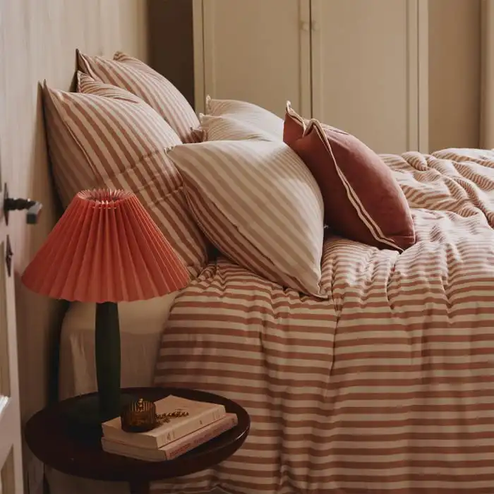 Cozy bedroom with striped bedding, a stack of books, and a trendy pink lamp on a bedside table, providing a warm, stylish ambiance.