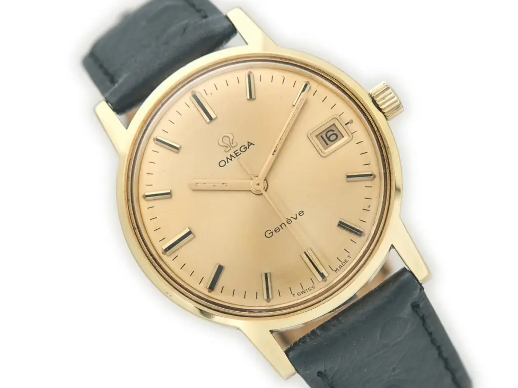Vintage Omega Genève watch with a gold dial, black leather strap, and date display at 3 o'clock position. Classic and elegant design.