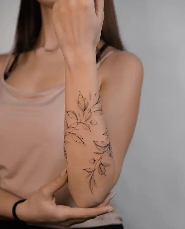 Elegant floral tattoo design on a woman's forearm, showcasing intricate black ink branches and leaves for a stylish, modern look.