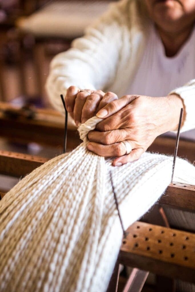 Our Artisans Hands weaving white wool yarn on a traditional loom, highlighting craftsmanship and textile artistry. | Sky Rye Design Hands weaving white wool yarn on a traditional loom, highlighting craftsmanship and textile artistry.