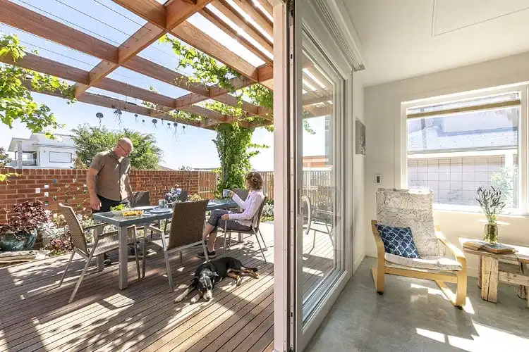 Sunlit patio with pergola, two people dining, and a dog resting, viewed from a bright interior room with a cozy chair.