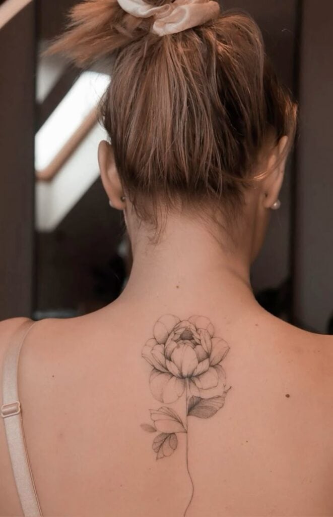 Woman with hair in a bun shows off an intricate flower tattoo on her upper back, featuring a delicate bloom and leaves.