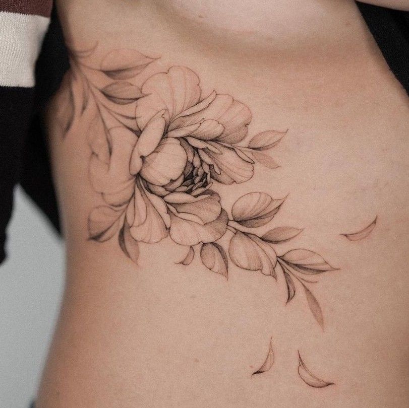 Delicate floral tattoo design on side of ribcage, featuring detailed petals and leaves in black and gray shading.